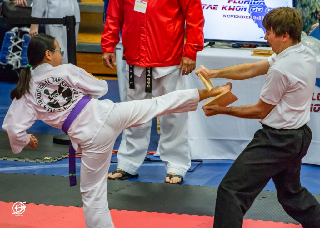 side view of tae kwon do girl kicking and breaking a board held by a young man
